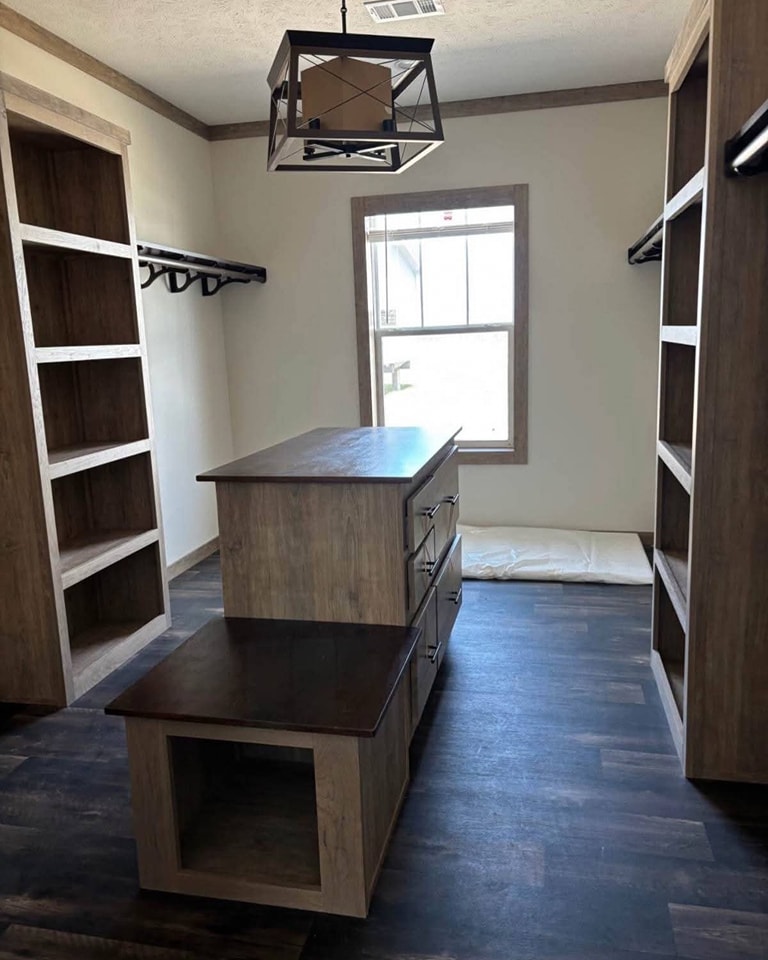 Spacious walk-in closet with dark wood floors, light wood cabinets, and open shelves. A modern pendant light hangs above a central island dresser.