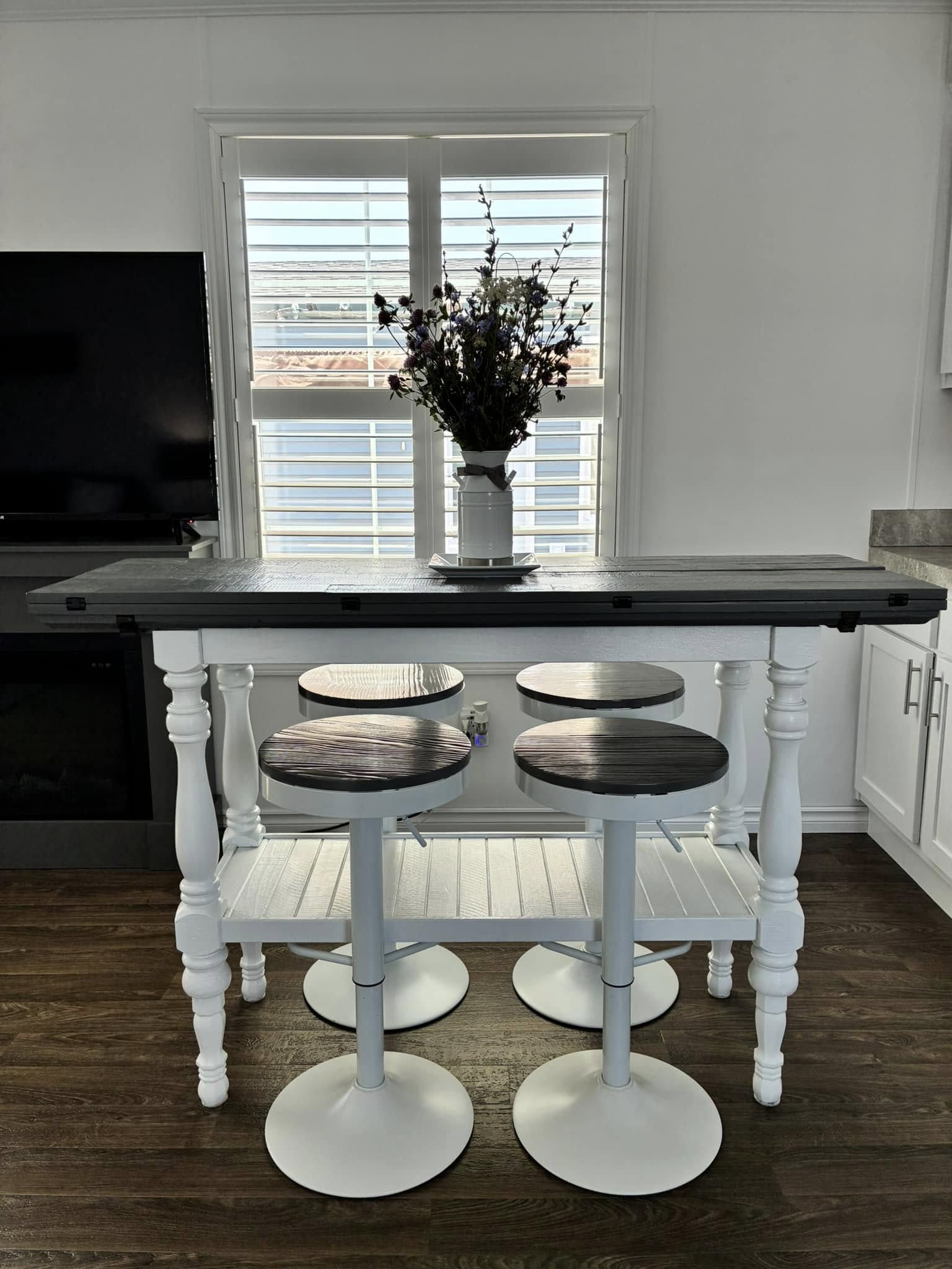 A modern kitchen features a tall, dark table with four sleek bar stools. A vase with vibrant purple flowers is illuminated by sunlight through shuttered windows.