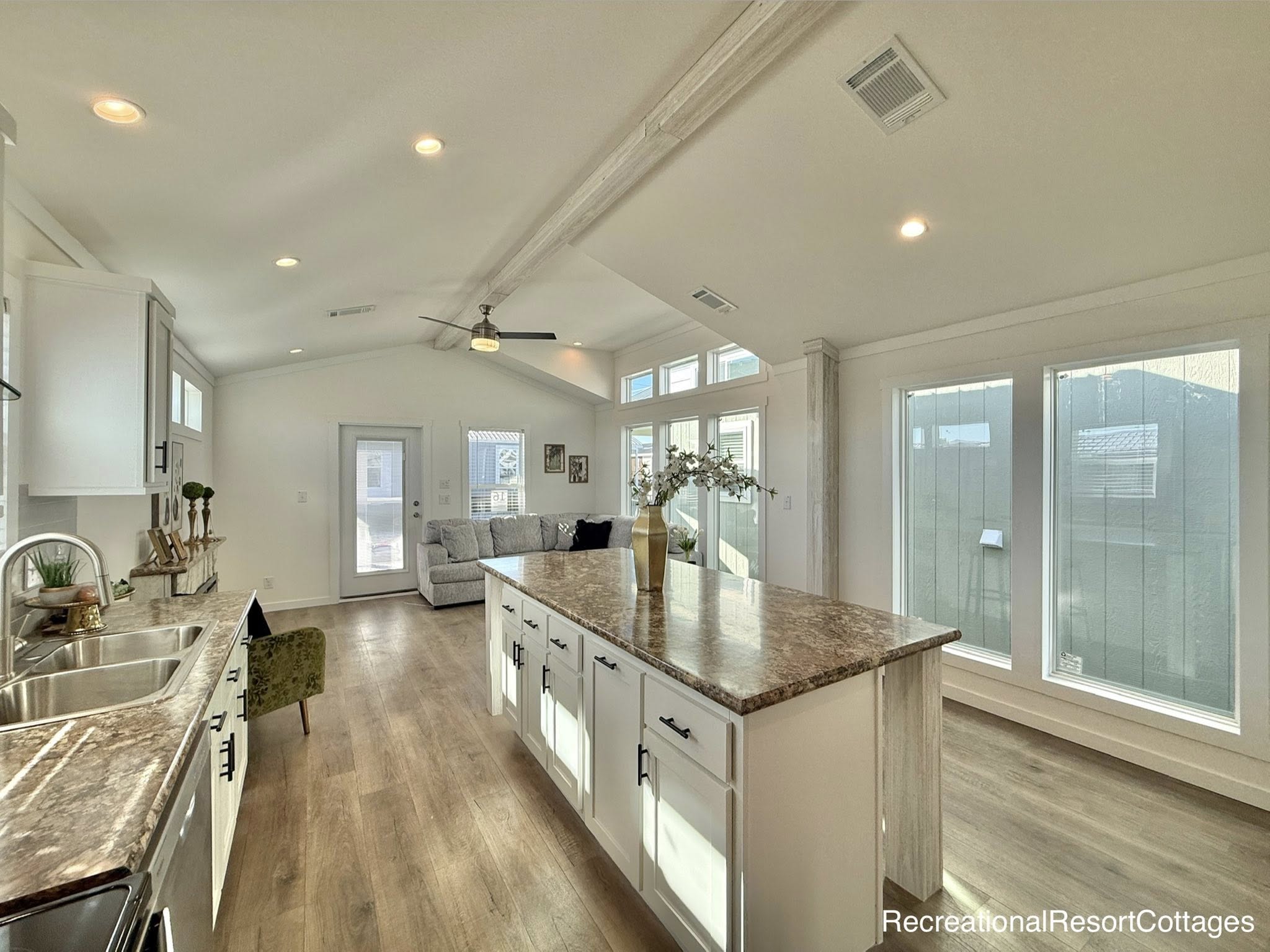 Bright, modern kitchen and living area with high ceilings. Features white cabinets, marble countertops, wooden floors, and a ceiling fan. Large windows offer natural light.