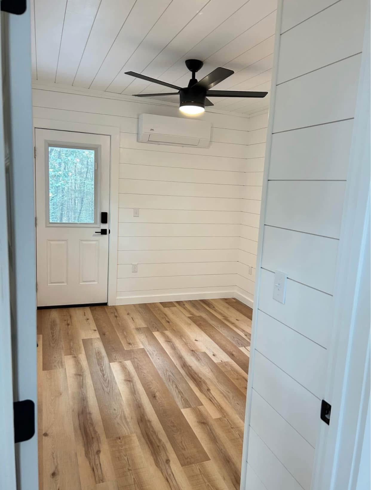 Cozy room with white shiplap walls, wood floor, and a black ceiling fan. A door with a window lets in natural light, creating a fresh, inviting feel.