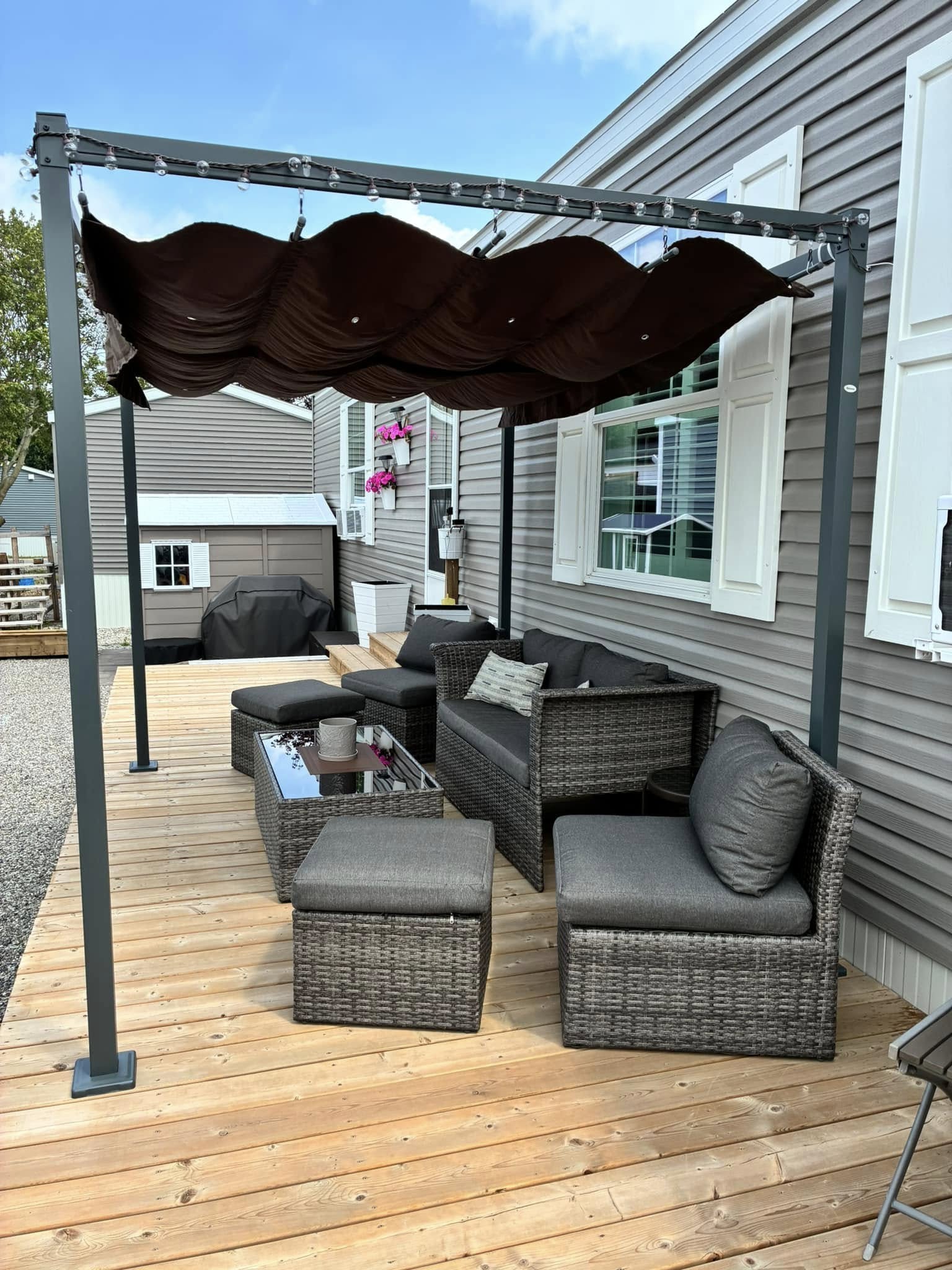 Outdoor patio with wicker furniture under a dark fabric pergola. Cozy cushions and a glass-top table add comfort beside a gray house with pink flowers.