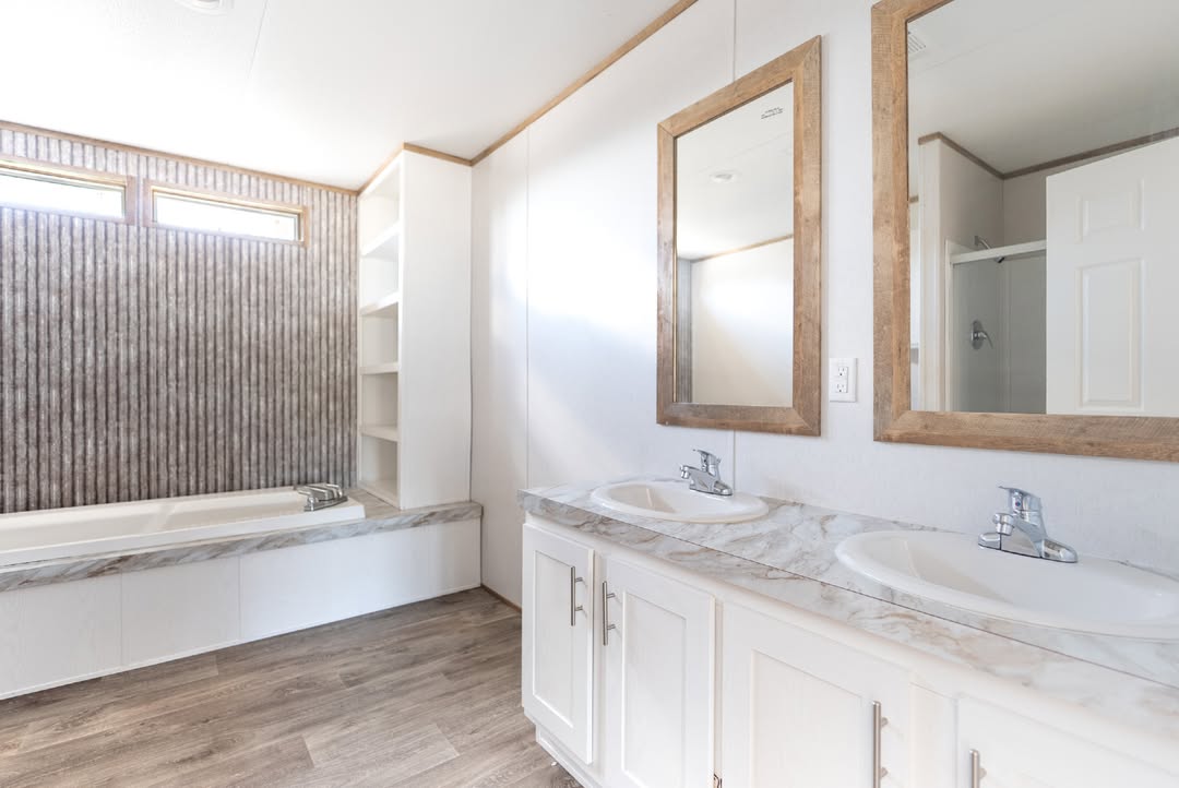 Modern, bright bathroom with wood accents. Features dual sinks with marble countertops, two mirrors, a bathtub with tile surround, and wooden flooring.
