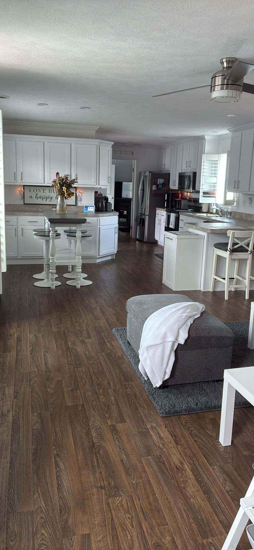 A modern kitchen with white cabinets, a marble countertop, and bar stools. Wooden floor, cozy gray ottoman with a throw, and a ceiling fan create a warm ambiance.