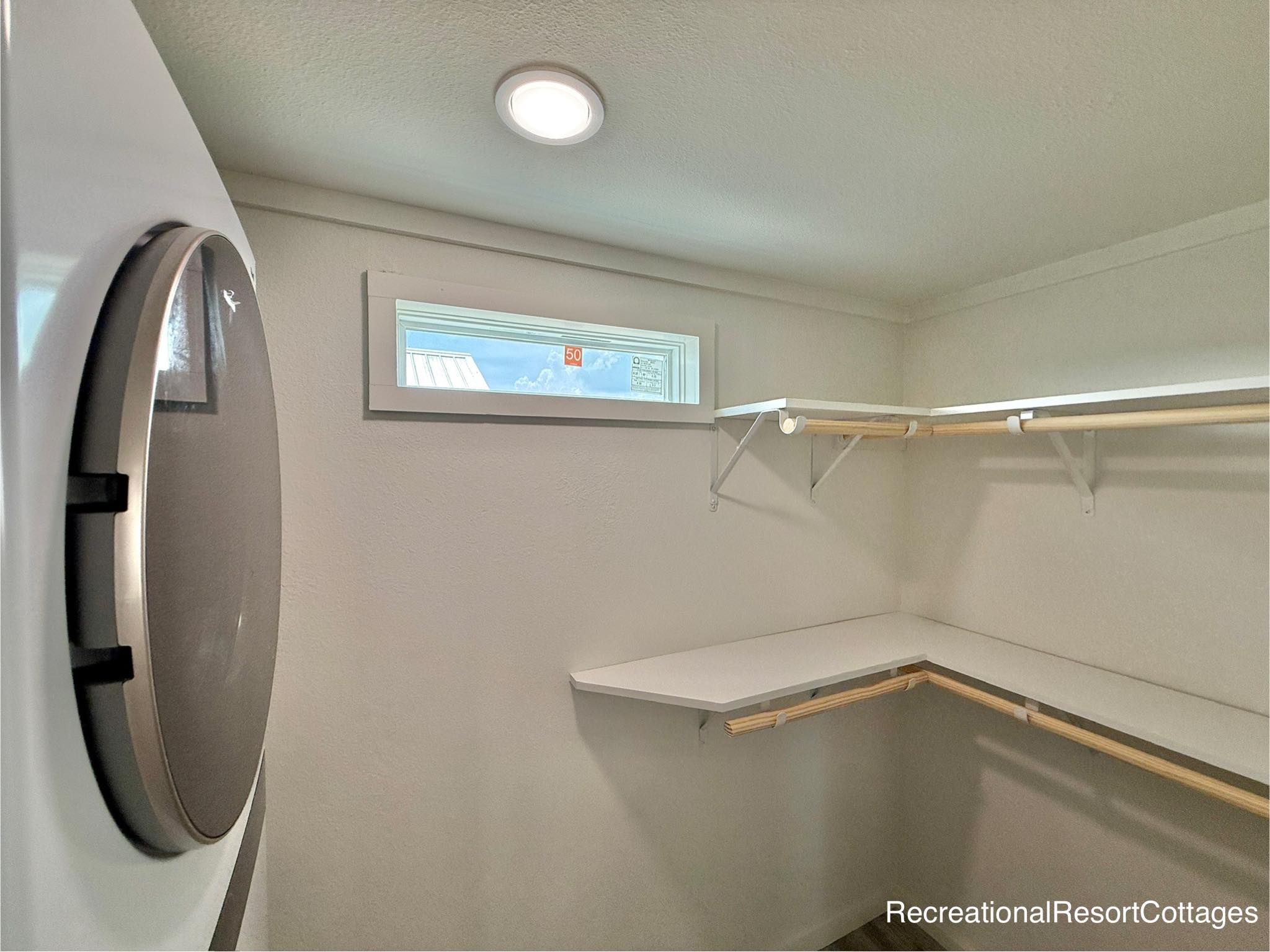 Laundry room with stacked washer and dryer, small window above, L-shaped white countertop, and shelving, creating a clean, minimalistic space.