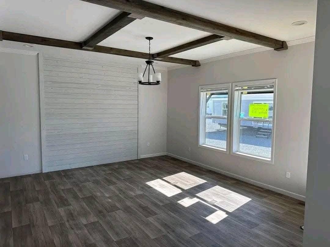 Spacious, empty room with gray wood flooring, white shiplap accent wall, and large windows. Exposed beams and modern light fixture add rustic charm.