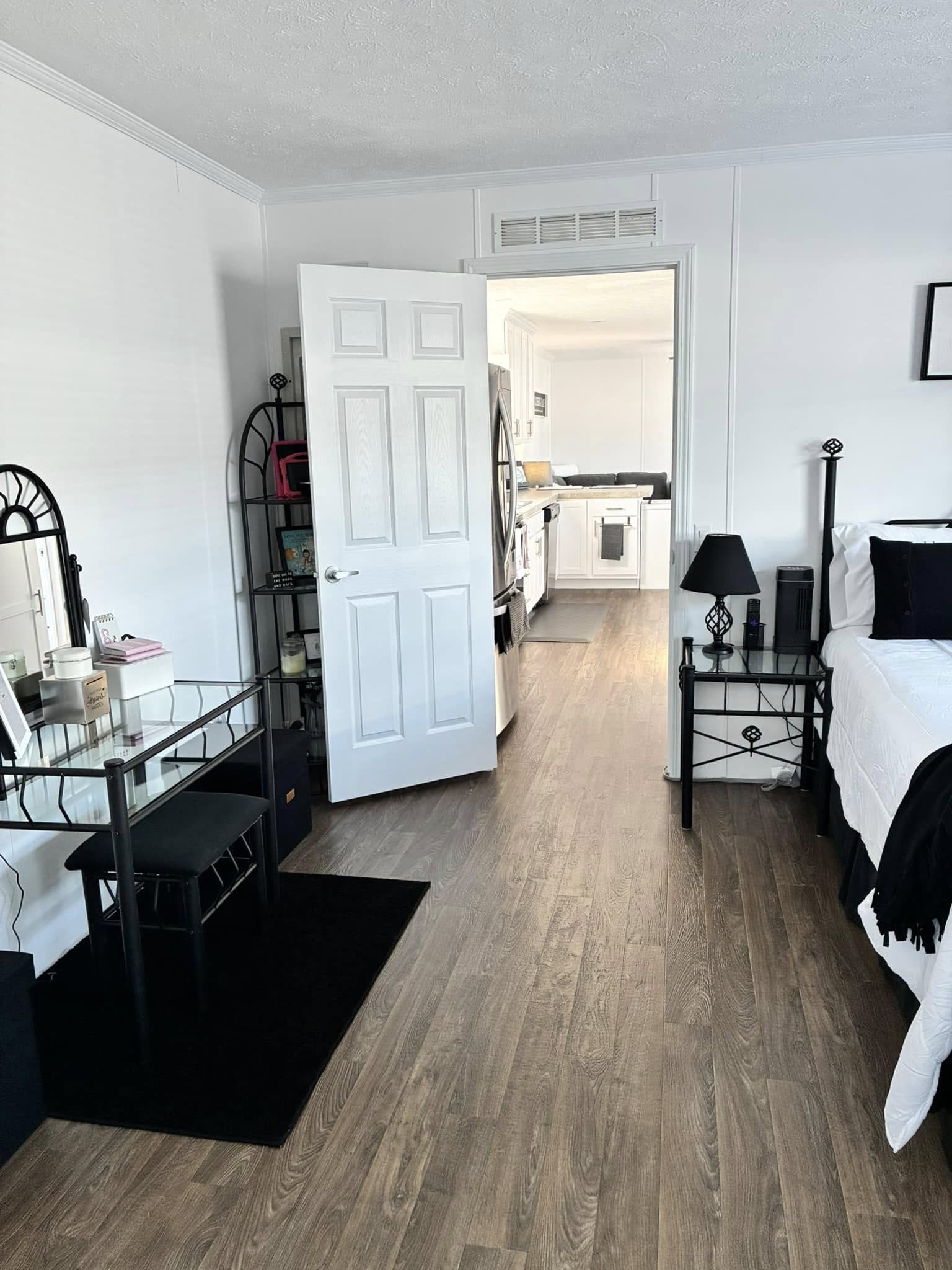 Bright room with wooden floors, a bed, and a glass desk with a chair. An open door reveals a modern kitchen. The ambiance is clean and cozy.