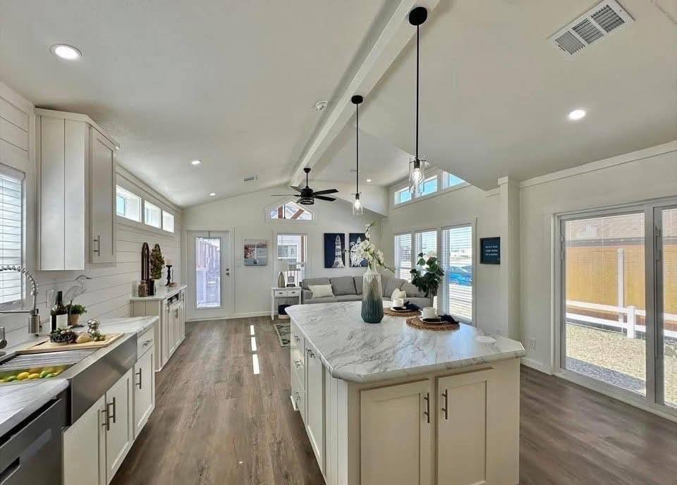 Bright, modern kitchen and living area with white cabinets, marble island, and wood flooring. Large windows and pendant lights add an airy feel.