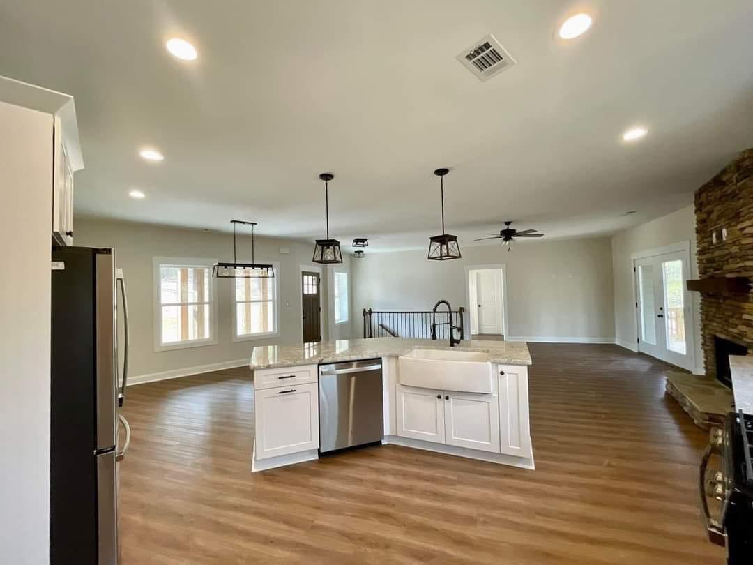 Spacious open-concept kitchen and living area with hardwood floors. Features a white island, hanging lights, stone fireplace, and large windows. Calm, inviting ambiance.