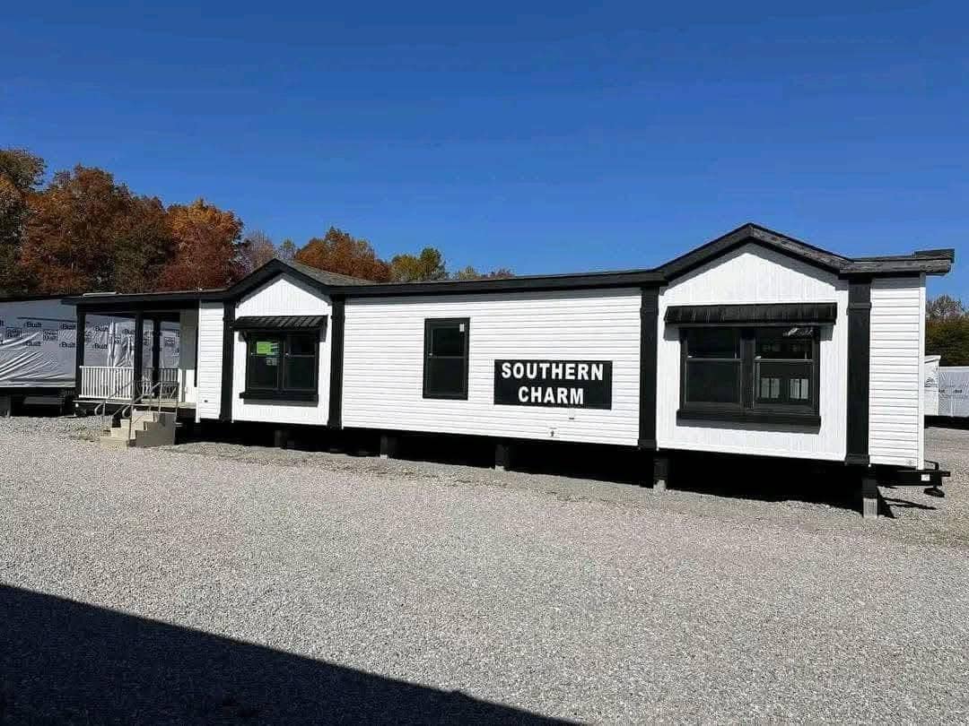 White modular home labeled "Southern Charm" with black trim and large windows, situated on gravel with autumn trees in the background under a clear blue sky.