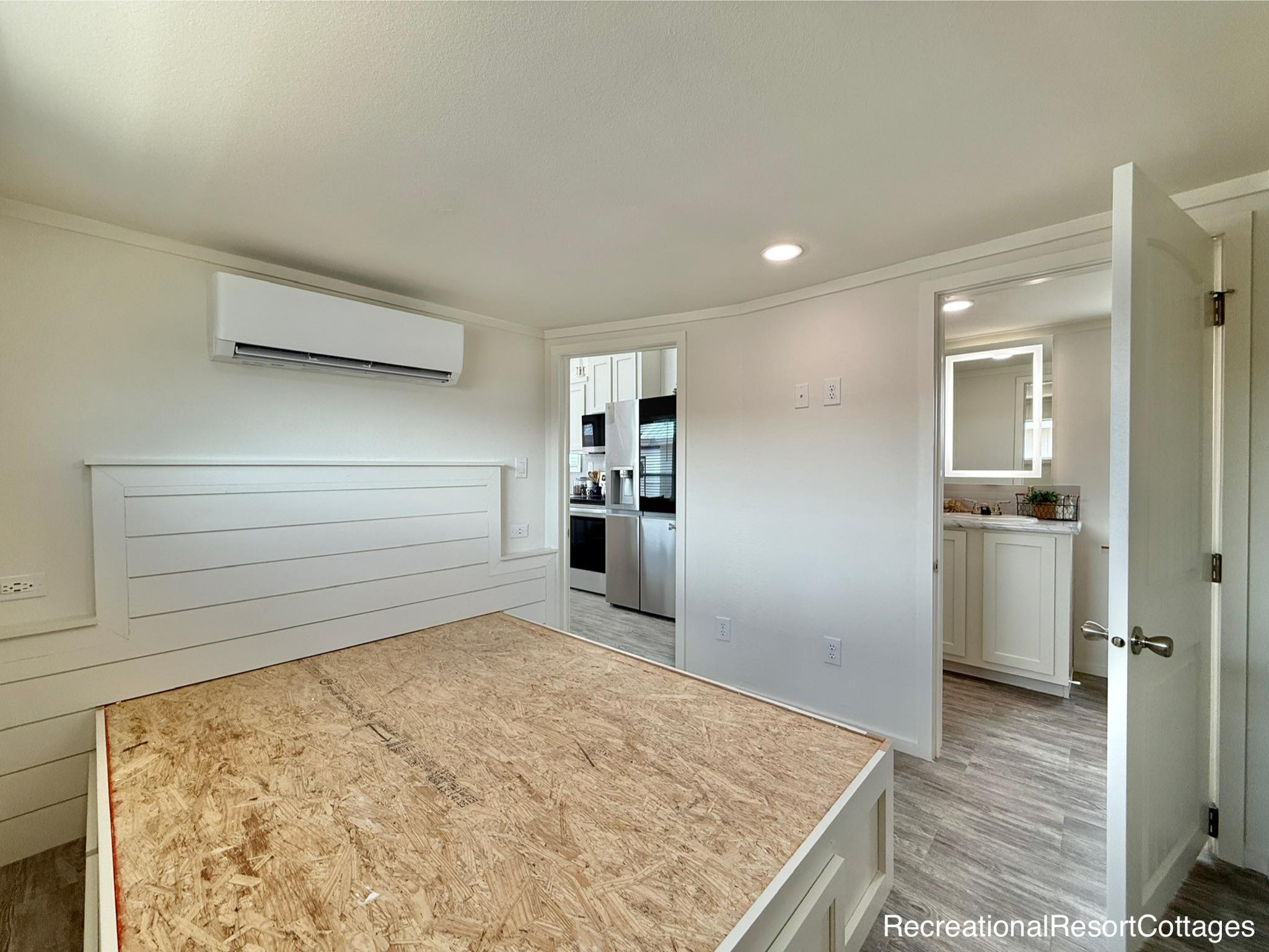 Minimalist white room with an unfinished wooden platform bed. Air conditioner on wall, open door shows a kitchen with stainless steel appliances, and a bathroom vanity visible. Bright, clean, modern feel.