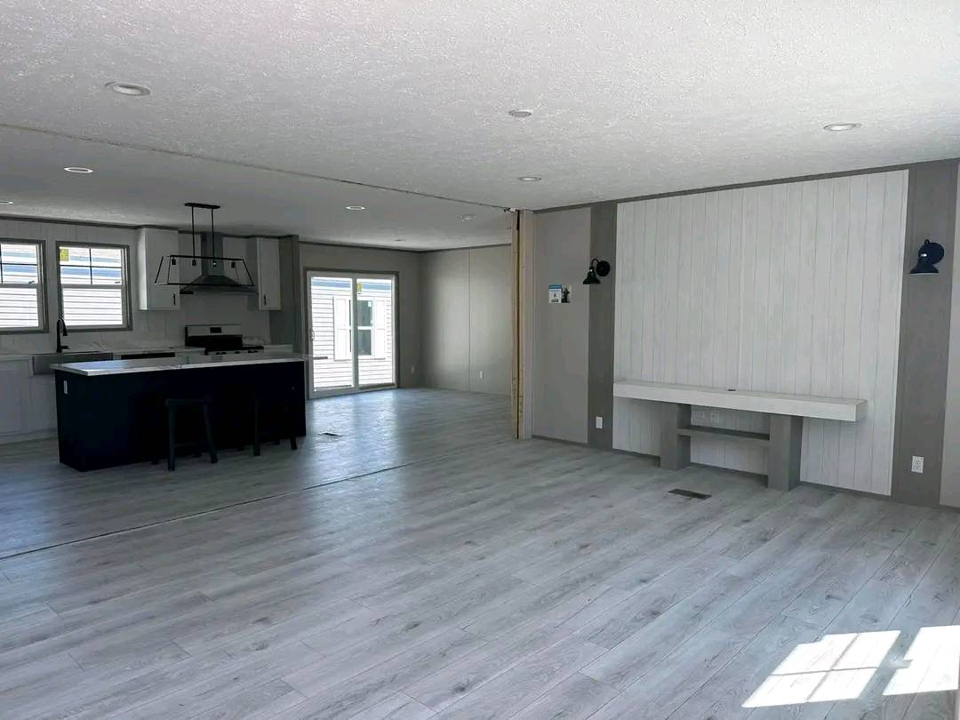 Spacious, modern open-concept room with light wood flooring, gray walls, a kitchen island, ceiling lights, and large windows letting in natural light.