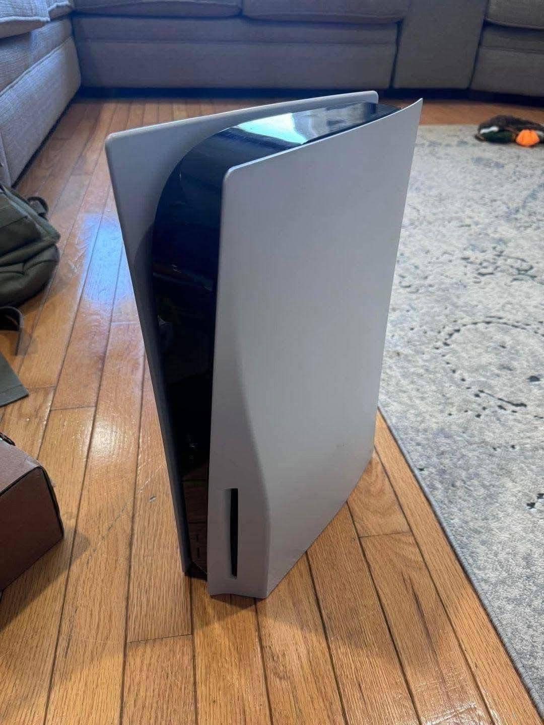 A PS5 console stands on a wooden floor beside a gray carpet in a living room. The sleek design is prominent, with beige couches and a bag in the background.