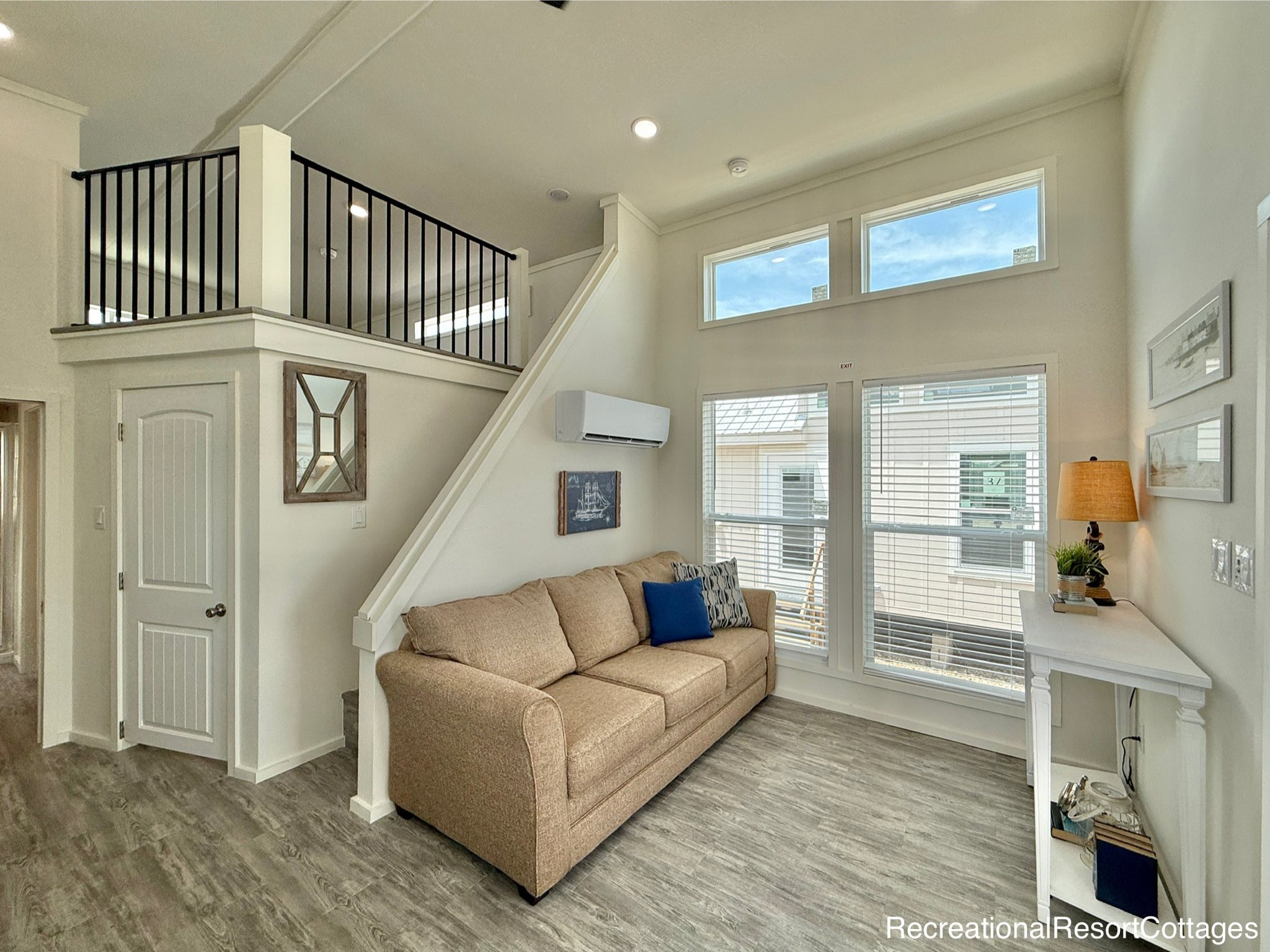 Bright, modern living area with high ceilings, a beige couch, and decorative pillows. Large windows, a loft with black railing, and a cozy atmosphere.