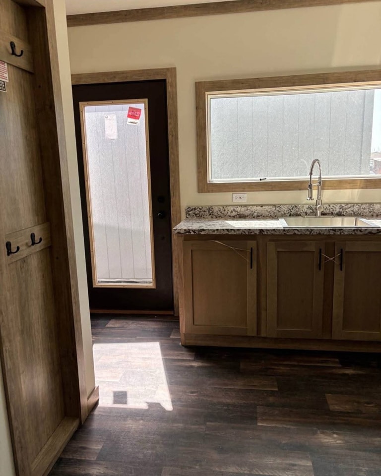 Small, bright kitchen with a large window above a sink on a marble countertop. Wooden cabinets and door, with hooks on the wall. Sunlight casts on dark wood floor.
