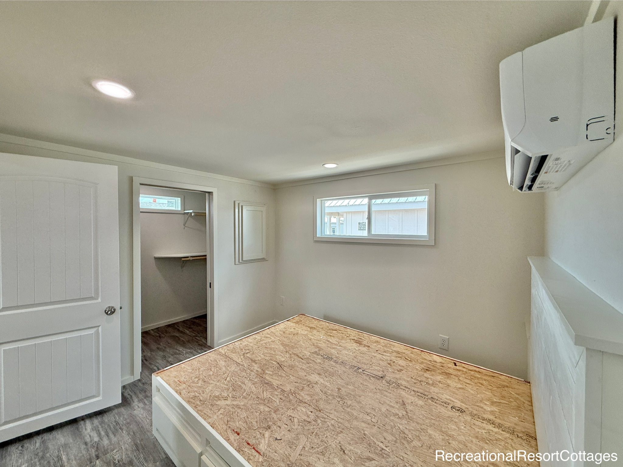 Minimalist bedroom with an unmade bed frame, white walls, and a small window. A door leads to a walk-in closet. Ceiling light and wall-mounted AC unit.