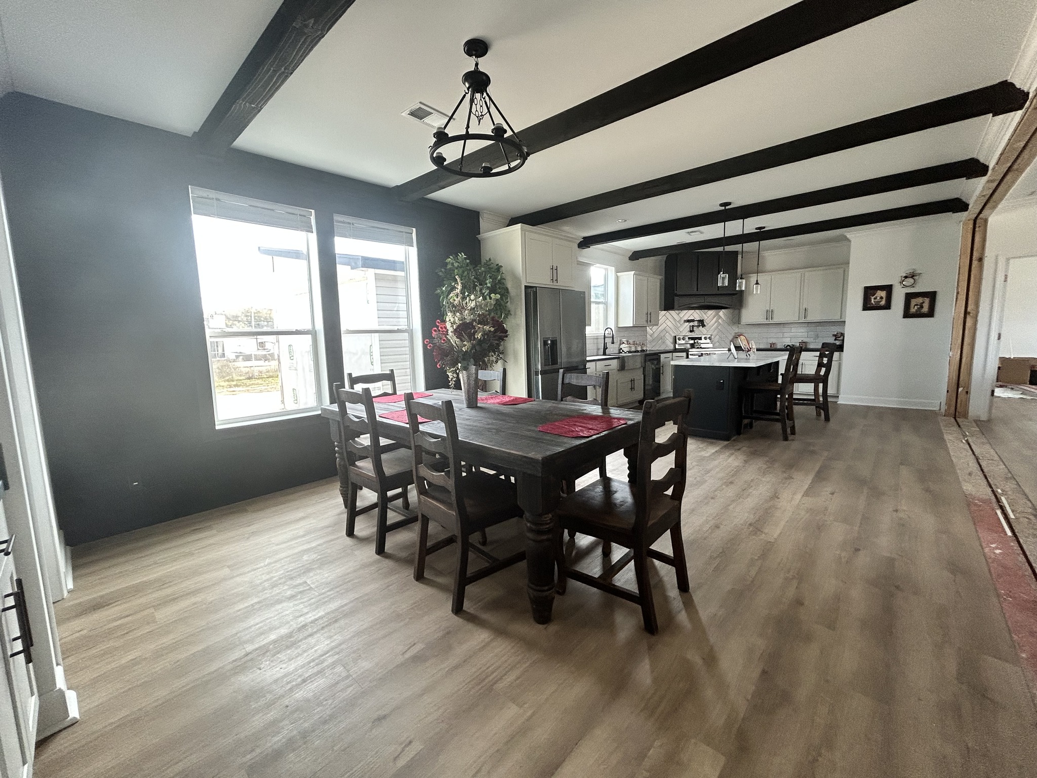 Modern dining room and kitchen with wood flooring. Dark wooden table, red placemats, and chairs centered. Black beam ceiling; sunny windows on left.