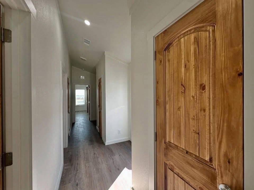 Narrow hallway with wooden door on the right, light wooden floor, and soft white walls. Sunlight filters through a distant window, creating a calm atmosphere.