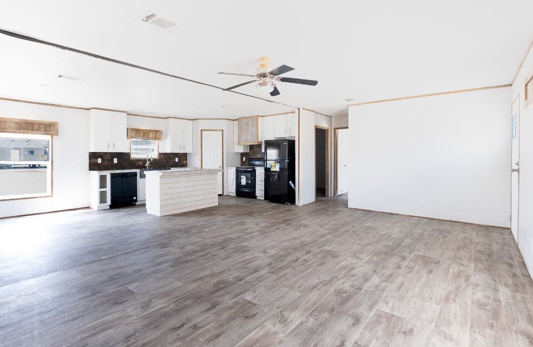 Spacious, empty room with light wood flooring, white walls, and a central kitchen area. Features a ceiling fan, black appliances, and large windows.