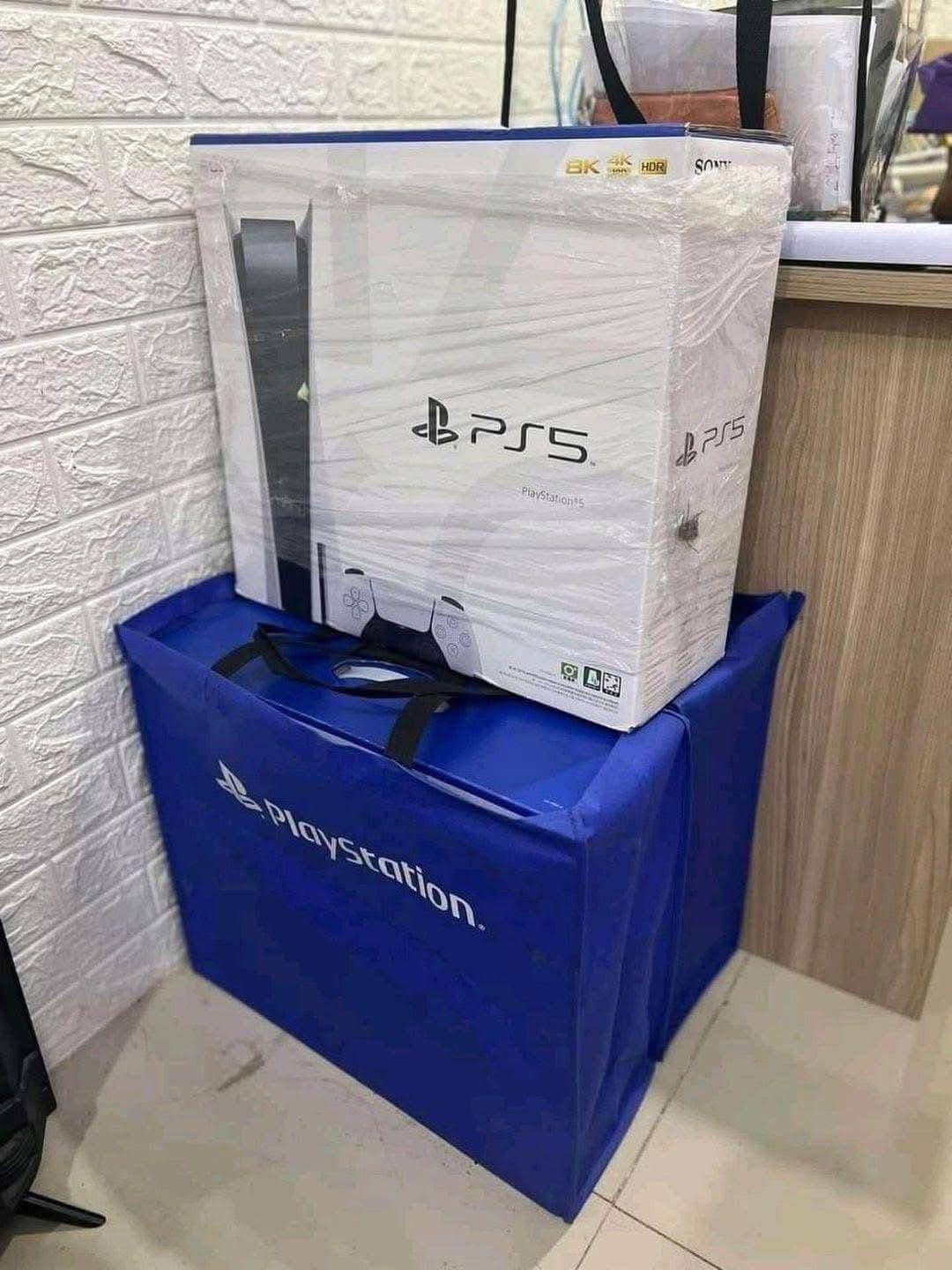 A boxed PlayStation 5 console is wrapped in plastic, resting on a large blue PlayStation-branded bag. The setting is indoors with a beige tiled floor.