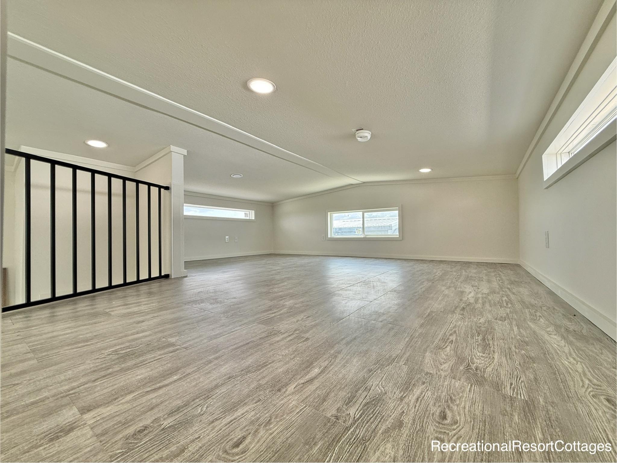 Spacious, minimalist loft with light wood flooring, a low ceiling, recessed lighting, and two small windows. The mood is bright and open.