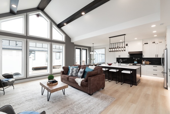 A modern open-concept living room and kitchen with vaulted ceilings and large windows. Features a brown sofa, wood coffee table, and sleek kitchen island.