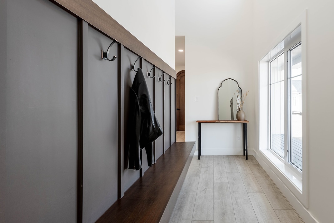 A modern entryway with a wooden bench and hooks on a gray paneled wall, a black jacket hanging. Opposite, a console table with a mirror and vase by a large window.