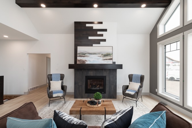 Modern living room with a high ceiling, centered around a black fireplace with abstract art. Two armchairs flank a wood coffee table. Large windows on the right.