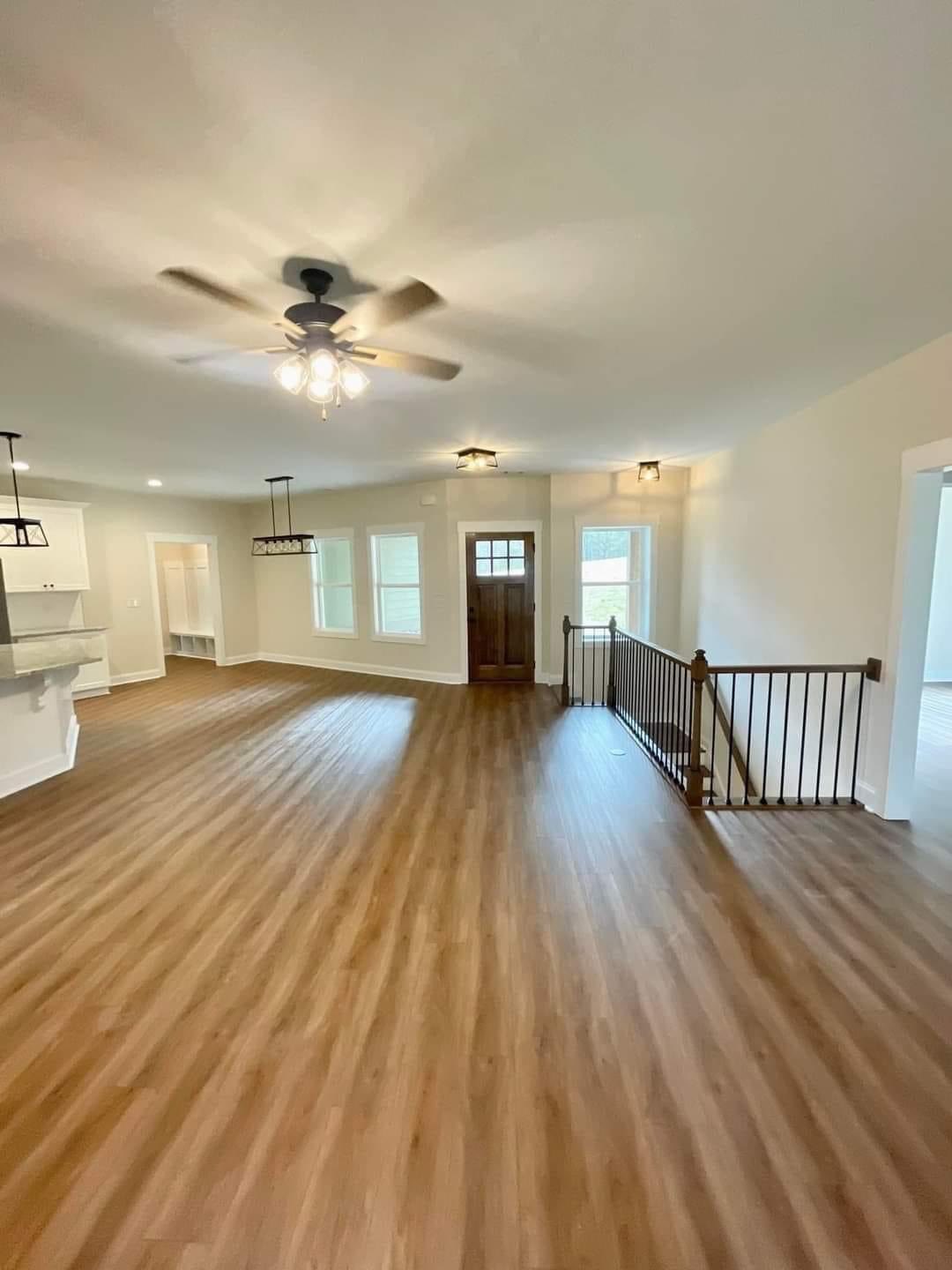 Spacious living area with wooden floors, beige walls, and ceiling fan. Features large windows, a wooden front door, and a staircase with a railing. Cozy and inviting.