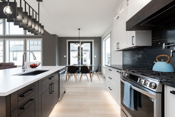 Modern kitchen with sleek, dark cabinets, white countertops, and stainless steel appliances. A blue kettle sits on the stove. Dining area with black chairs and a light wood floor is visible in the background. Abundant natural light and minimalist decor create an inviting, contemporary feel.