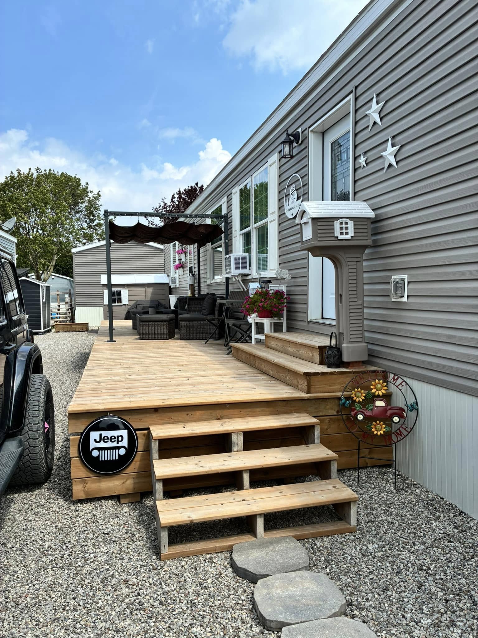 Wooden deck of a mobile home with steps, Jeep sign, and a decorative mailbox. Chairs, flowers, and star accents create a cozy, inviting vibe.