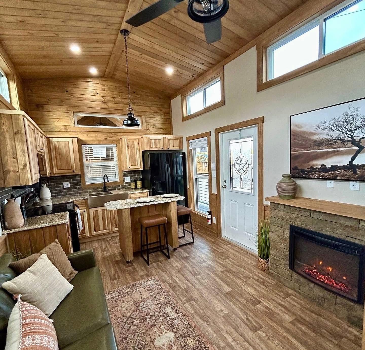 Cozy, rustic living room features wood-paneled walls and ceiling, stone fireplace, green sofa, and kitchen with island, creating a warm, inviting atmosphere.