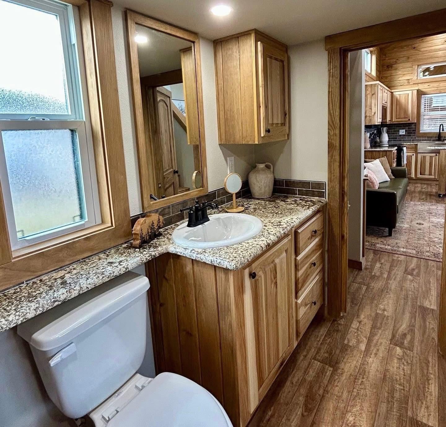Cozy bathroom with wood accents, featuring a granite countertop, sink, mirror, and toilet. Doorway leads to a rustic kitchen and living area.