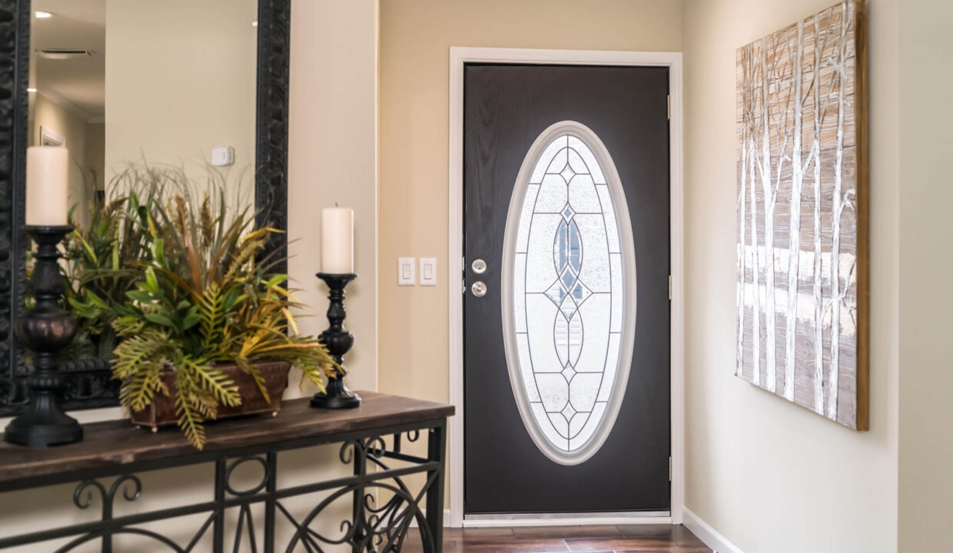A hallway with a decorative oval glass-paneled front door. A table on the left holds plants and candles. A tree-themed artwork hangs on the right wall.