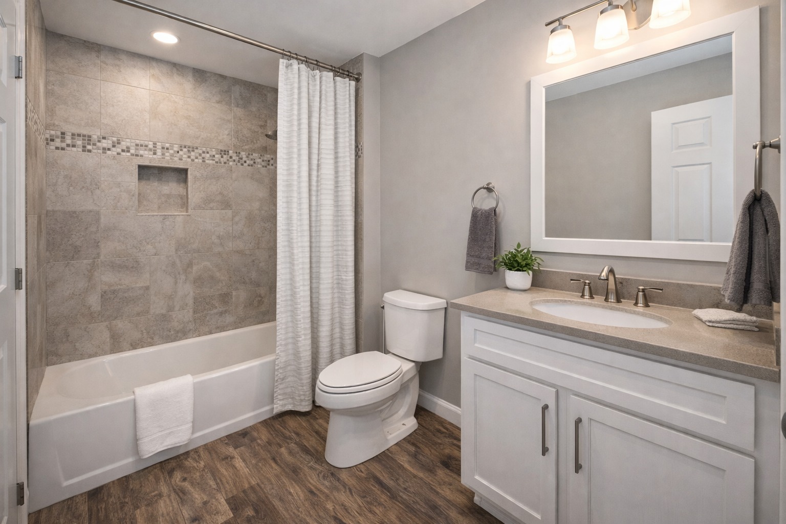 Modern bathroom with a bathtub and shower combo on the left, featuring beige tiles and a white curtain. A white vanity and sink with a mirror and soft lighting are on the right, accompanied by a small plant. The floor is dark wood, creating a clean and cozy ambiance.