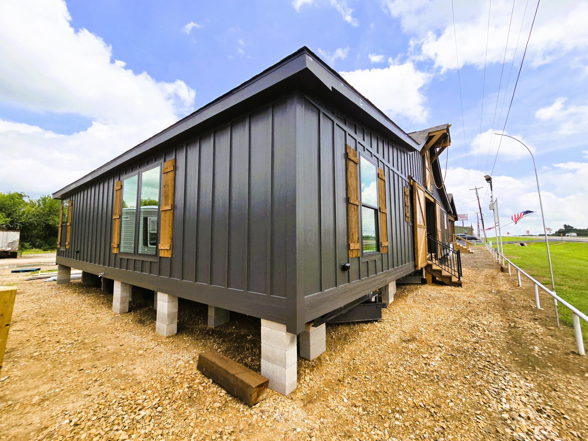 Modern tiny house on raised foundation with dark wood siding, large windows, and rustic shutters. Clear blue sky and American flag in the background.