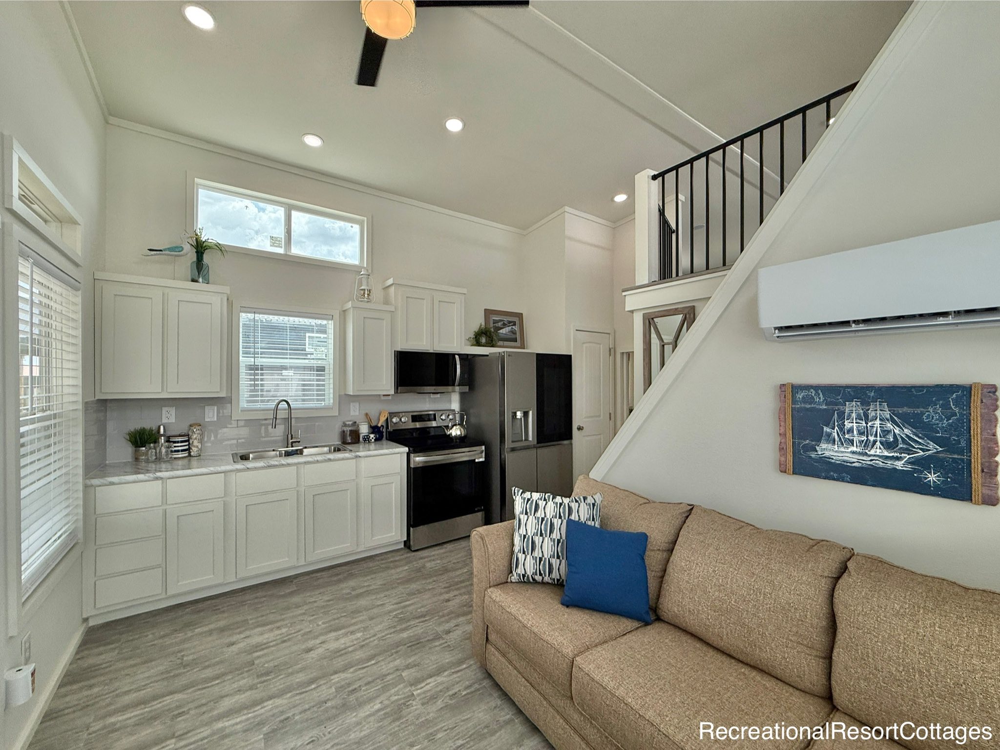 Modern, airy tiny home interior featuring a compact kitchen with white cabinets, stainless steel appliances, and a cozy living area with a beige sofa and nautical decor.