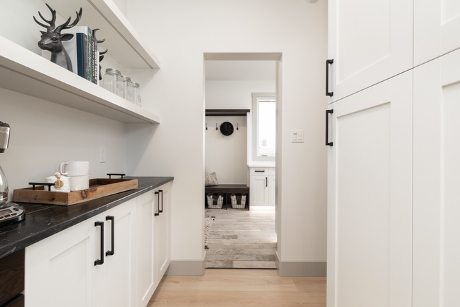 Narrow white pantry with black countertop and cabinets, featuring shelves with decor and glasses. Light wood floor leads to a bright mudroom.