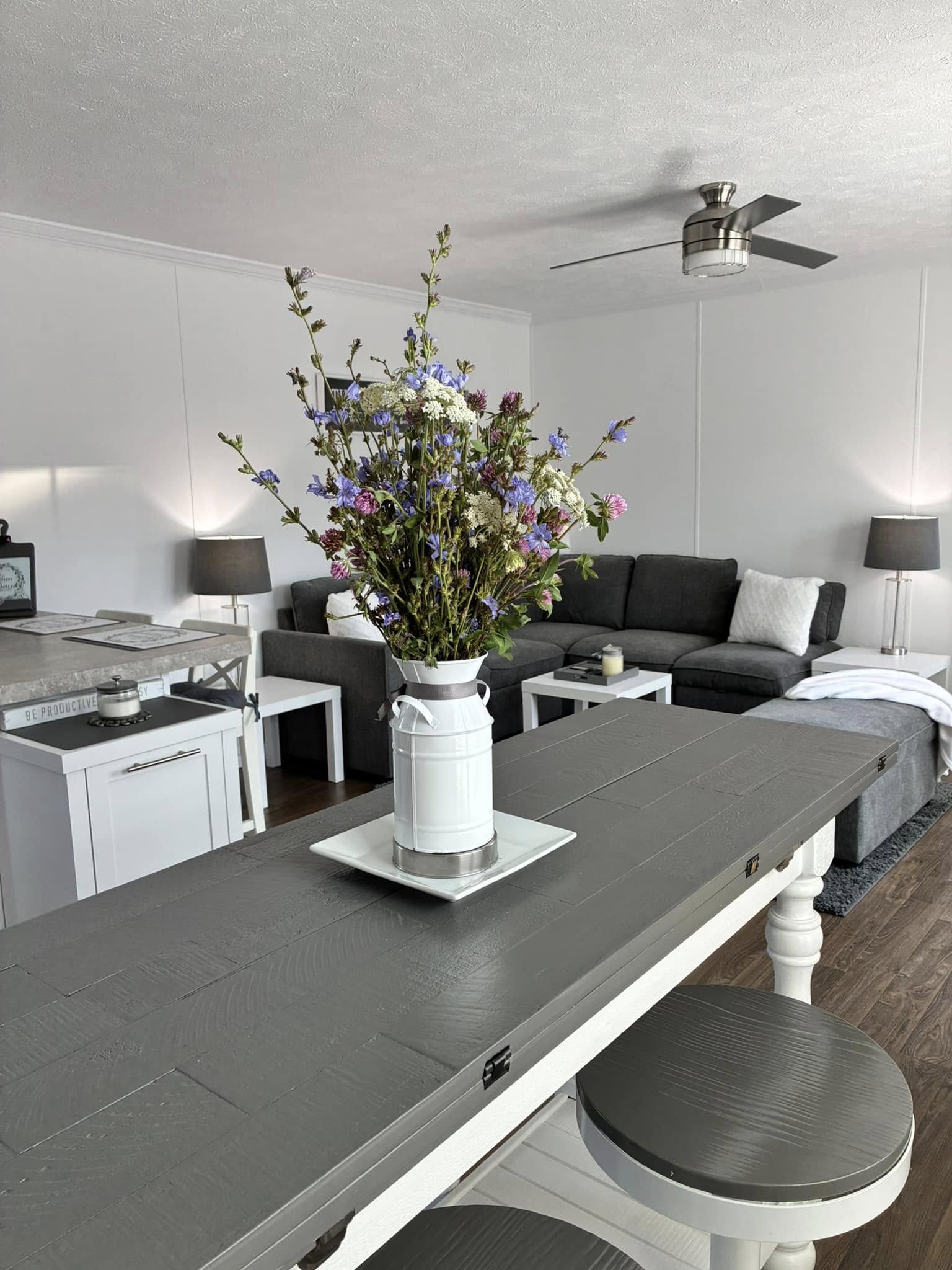 Modern living room with a dark gray sofa, white walls, and a ceiling fan. A large vase of colorful flowers sits on a gray table, creating a cozy ambiance.