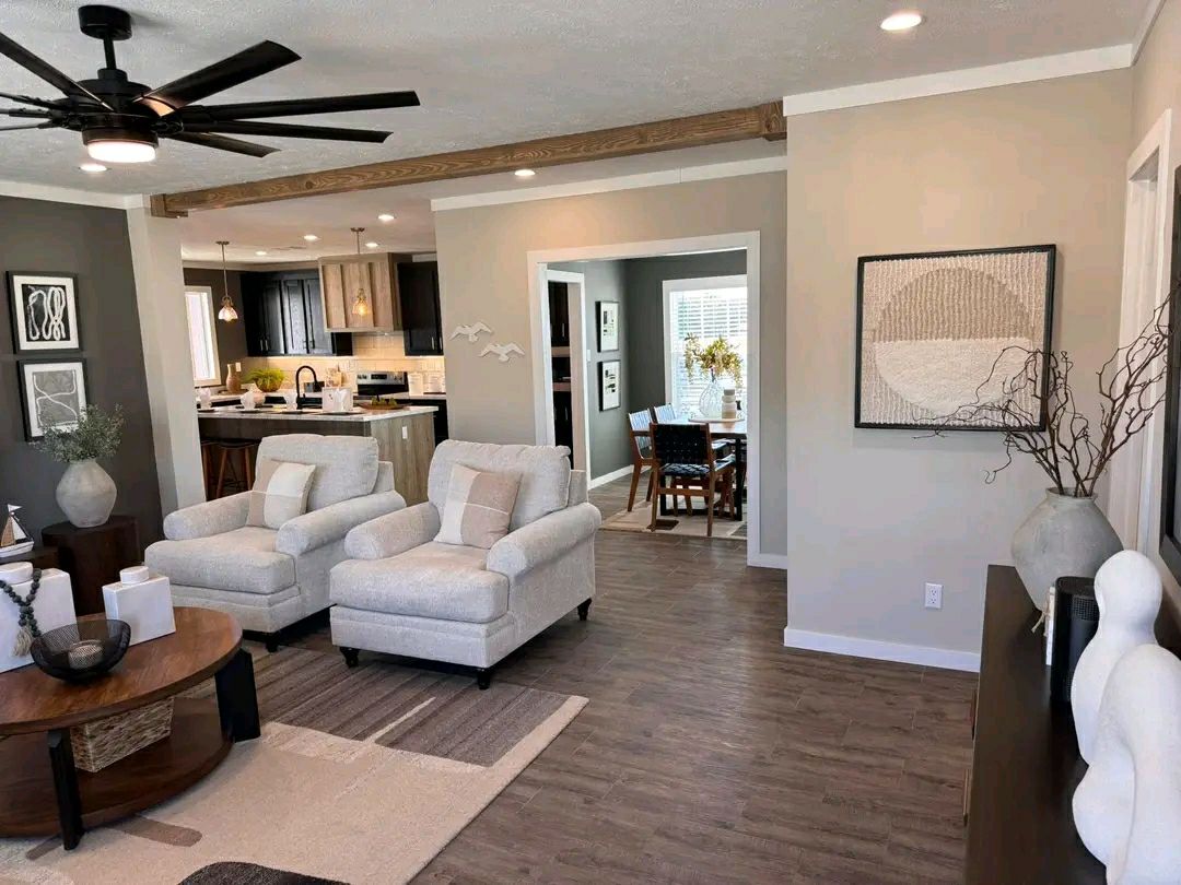 Modern living room with light gray armchairs, wood beams, and soft lighting. Open kitchen and dining area in the background. Neutral tones convey calmness.