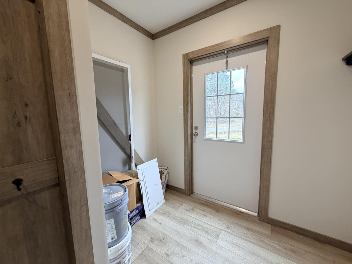 A small, tidy entryway with a wooden floor, beige walls, and a door with a window panel. Boxes and paint cans are stacked in a corner, creating a sense of organization.