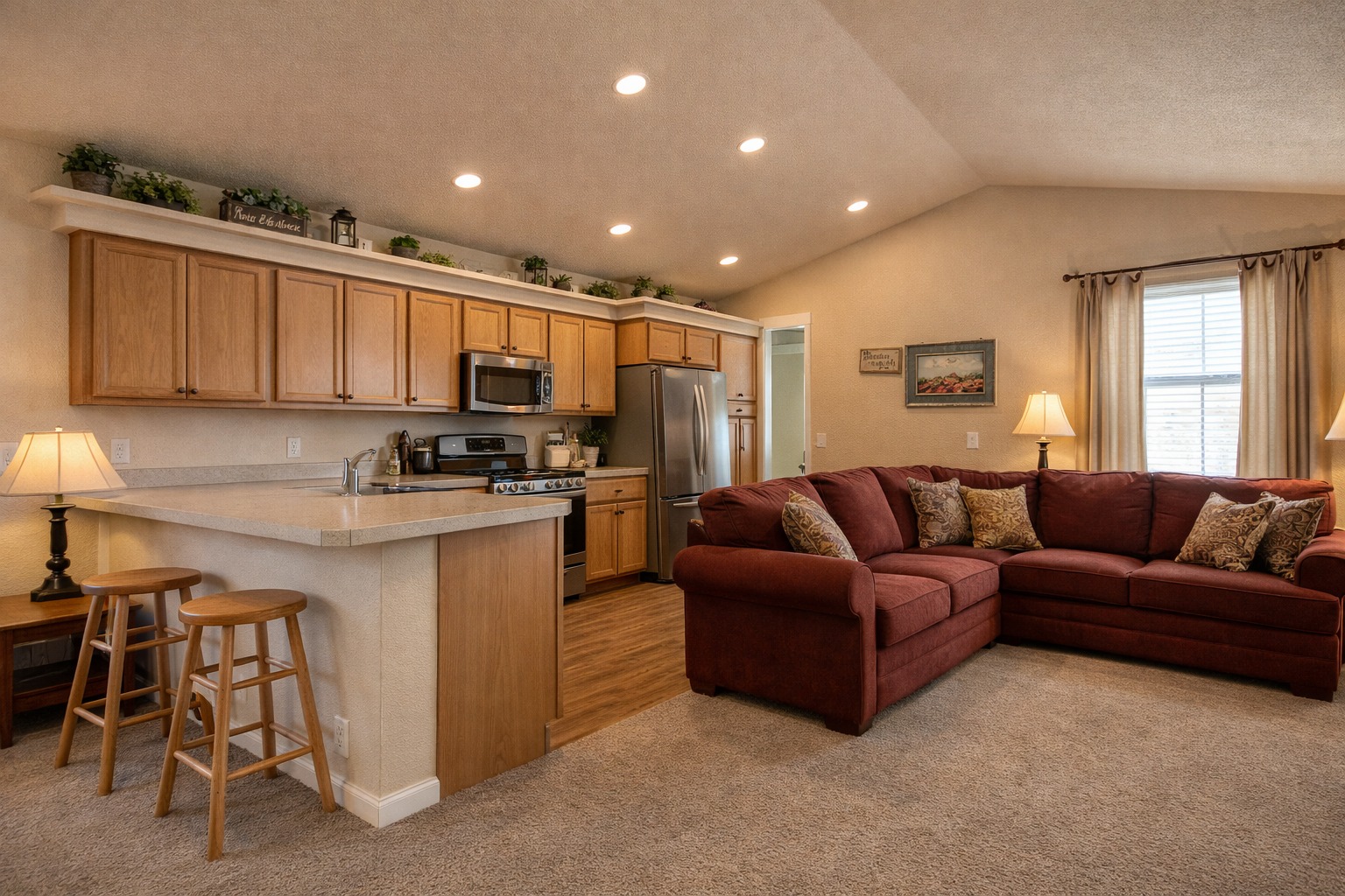 Warm, cozy living room and kitchen. Wooden cabinets, stainless steel appliances, red sectional sofa, soft lighting, and carpeted floor create an inviting atmosphere.