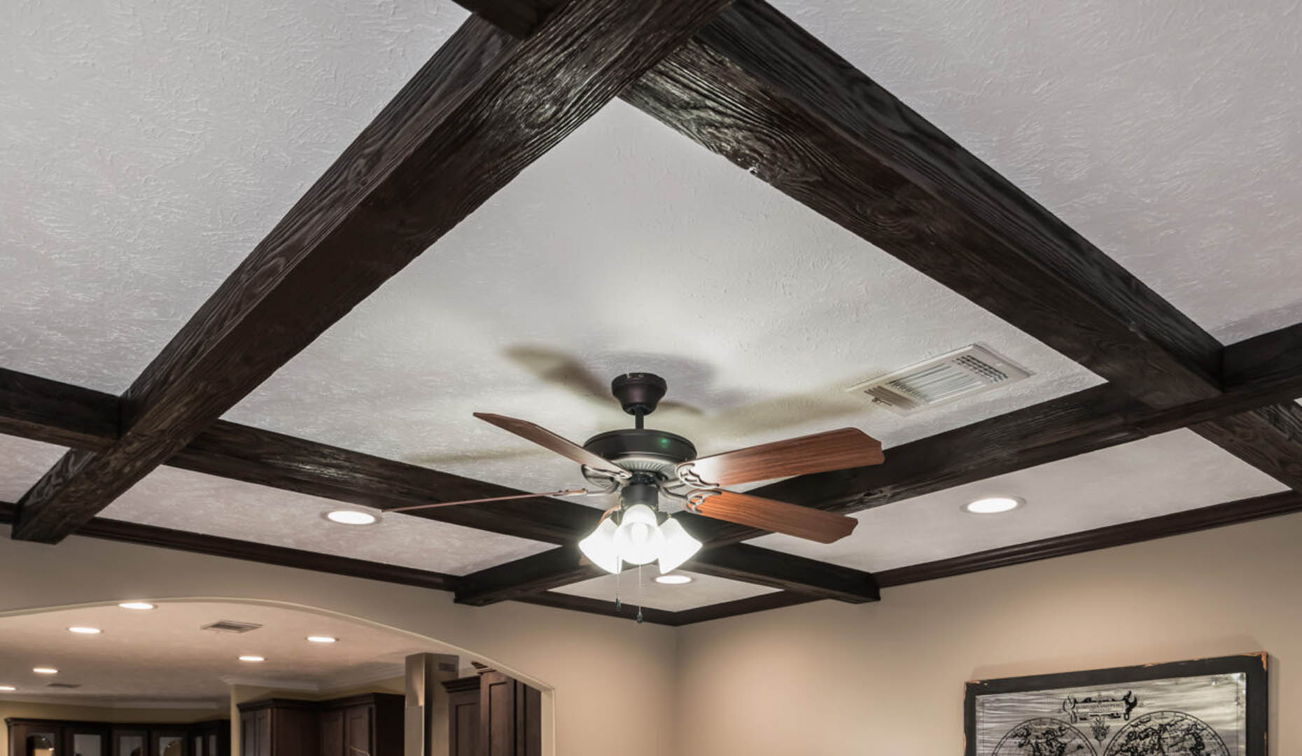 Ceiling with dark wooden beams forming a grid pattern, centered with a ceiling fan with wooden blades and lights. Room has recessed lighting and neutral-toned walls.