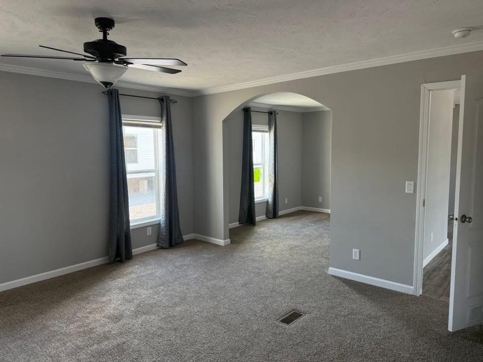 A spacious, empty living room with gray walls and carpet, featuring large windows with blue curtains. A ceiling fan hangs centrally, creating a calm, airy feel.