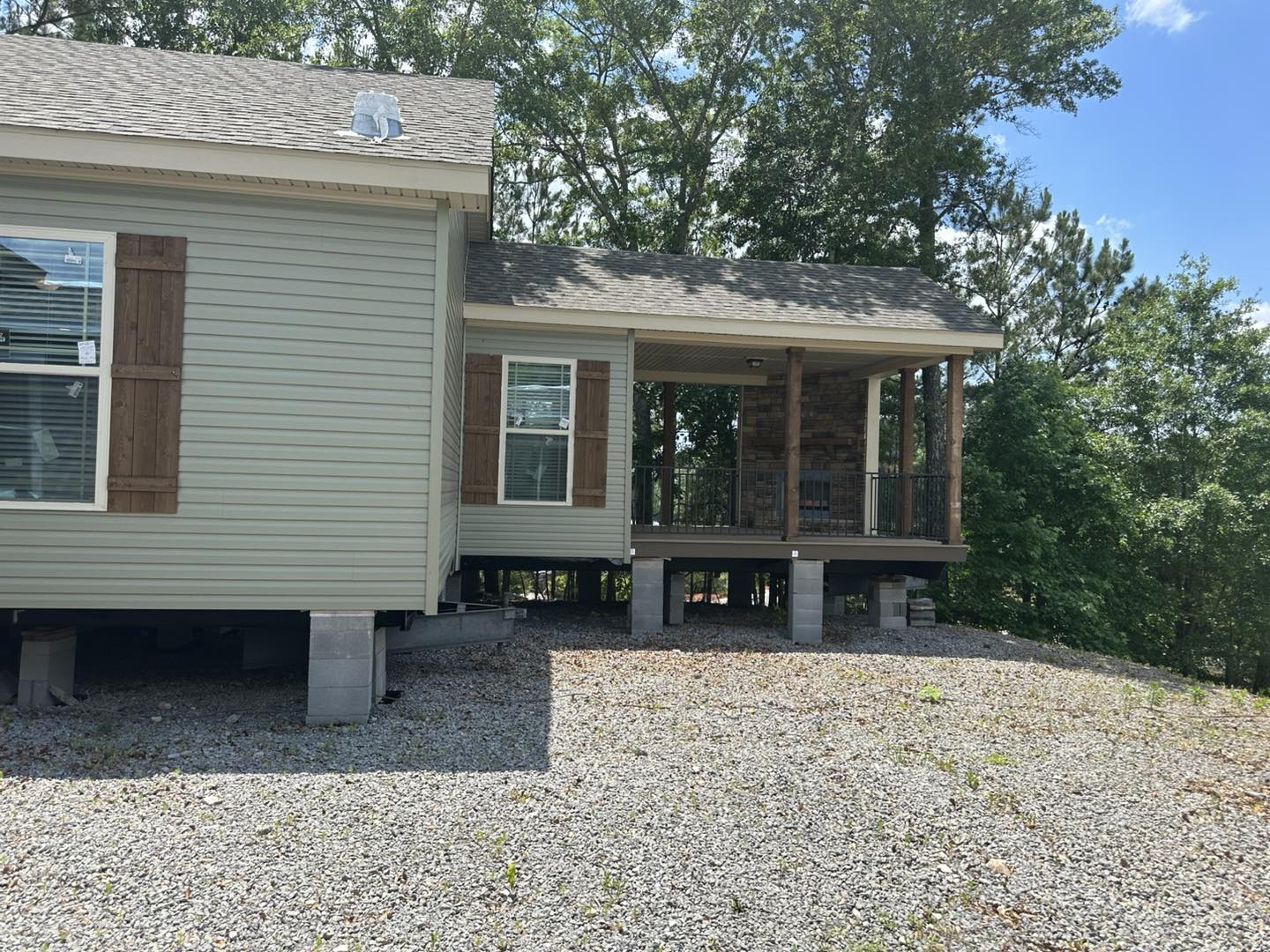 A beige manufactured home on a gravel lot, elevated on concrete blocks. It features wooden shutters and a covered porch with forest views. Daylight setting.