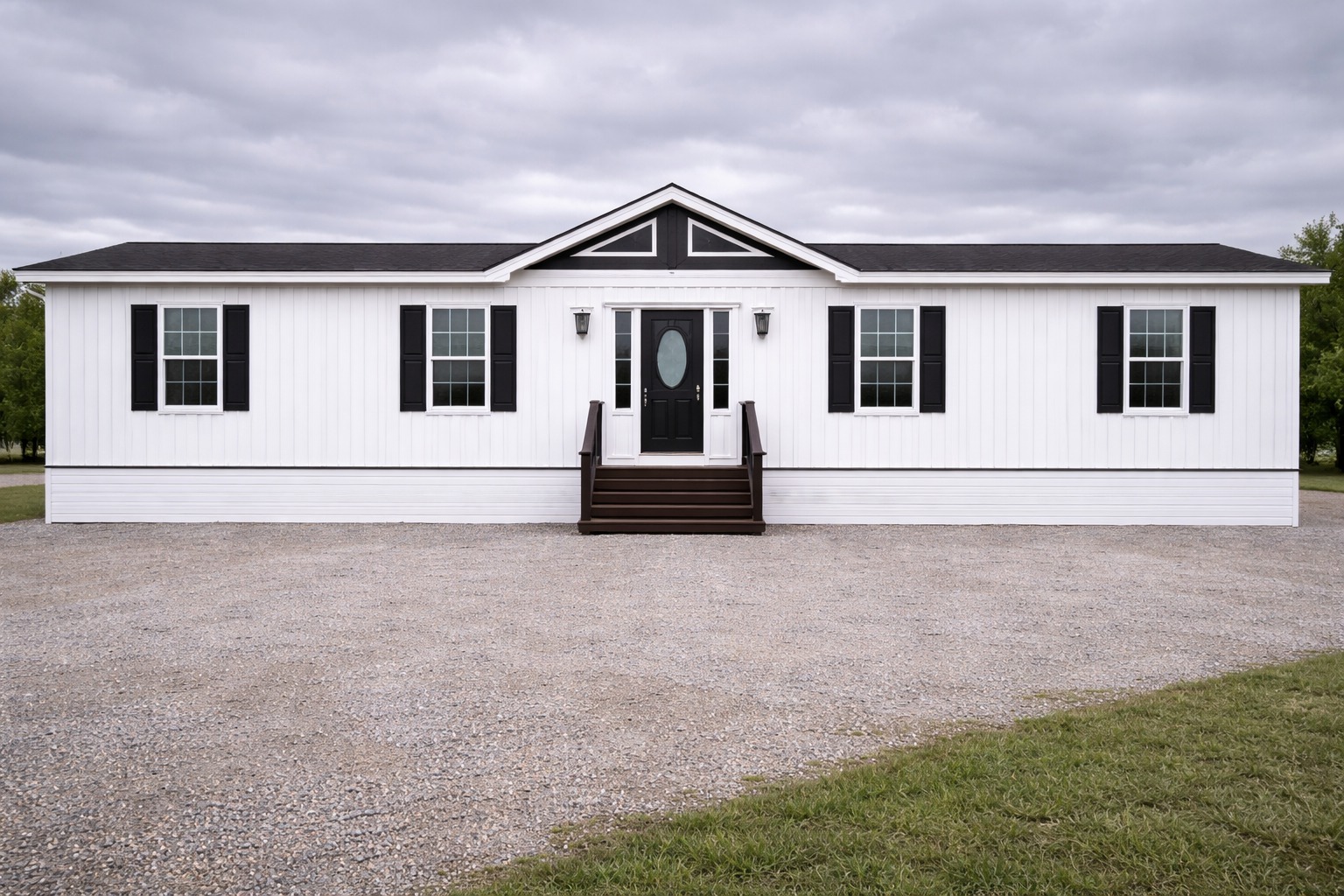 A white mobile home with black shutters and a centered dark door. It sits on a gravel driveway with cloudy skies overhead, evoking a calm rural setting.