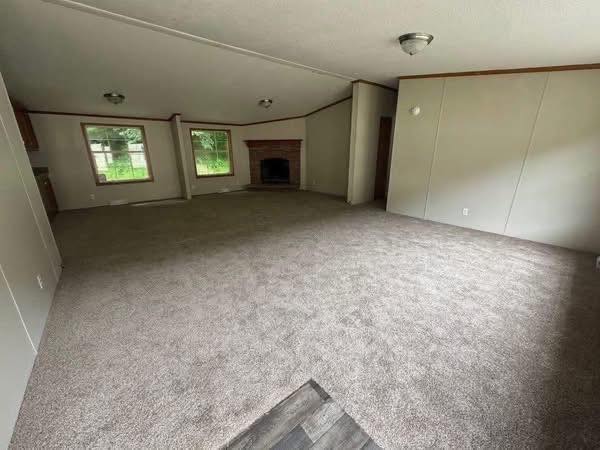 Spacious, empty living room with light gray carpet, beige walls, and two windows showing greenery outside. A brick fireplace adds a warm focal point.