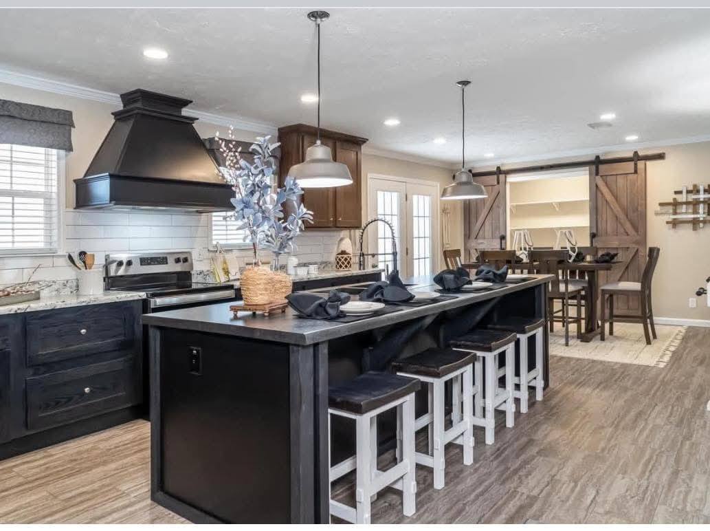 Modern kitchen with a large black island featuring four stools, pendant lighting, and a decorative vase. In the background, a dining area with barn doors and light wood flooring creates a cozy, inviting atmosphere.