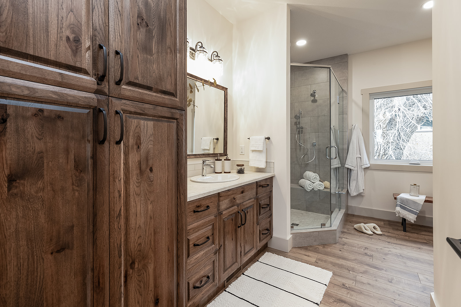 Rustic bathroom with wooden cabinets, double sink vanity, and glass shower. Mood is warm and cozy, with soft lighting and neutral tones.