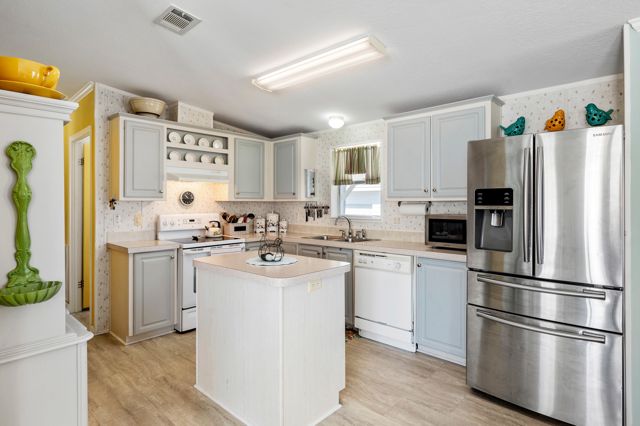 A cozy kitchen with light gray cabinets, a central island, and stainless steel fridge. Bright and inviting, the space has a warm, homely feel.