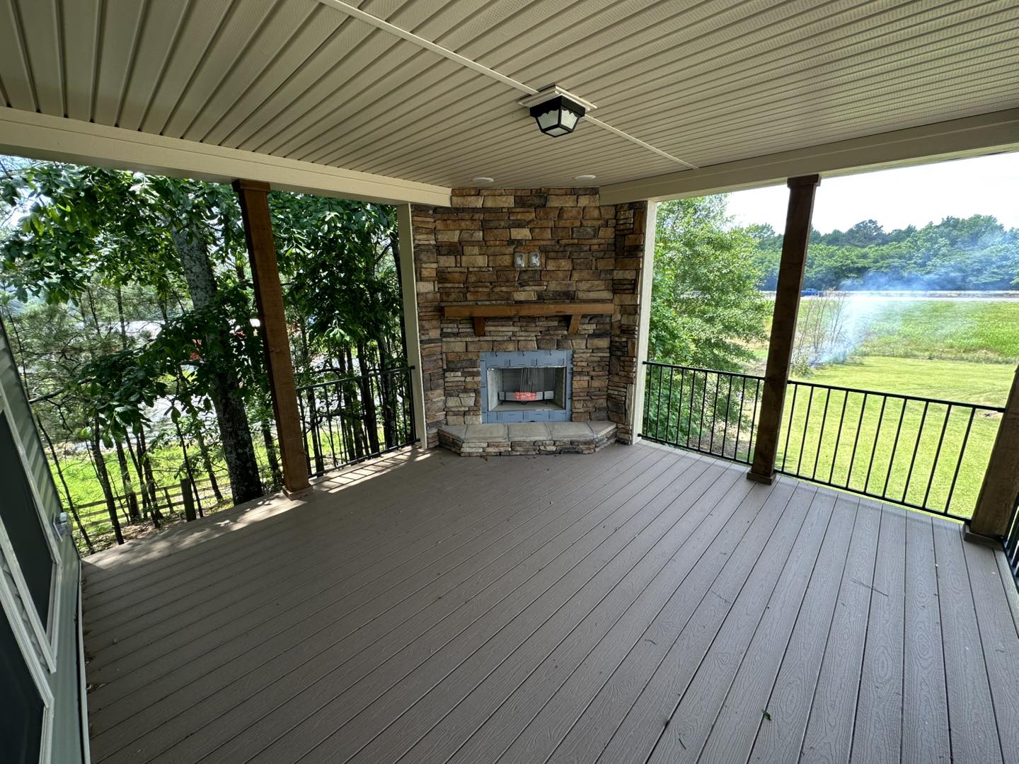 Covered outdoor patio with stone fireplace and wood ceiling. The patio overlooks lush greenery and a calm pond, creating a serene environment.